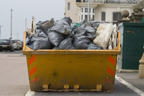 A large yellow industrial skip filled with black and white plastic rubbish bags, situated on a paved street in an urban setting. The skip's exterior shows signs of dirt and wear, with red reflective striping on the sides. Behind the skip, there is a row of parked cars and a glimpse of residential buildings, including a white multi-story structure with bay windows and decorative stonework. To the right, a green waste collection bin is partially visible, along with a stone bollard. The sky appears overcast, casting diffuse natural light over the scene. The image conveys a typical rubbish removal scenario in Hounslow, with the skip positioned on the street for waste collection by a commercial waste service such as Commercial Waste Hounslow, supporting local rubbish disposal needs.