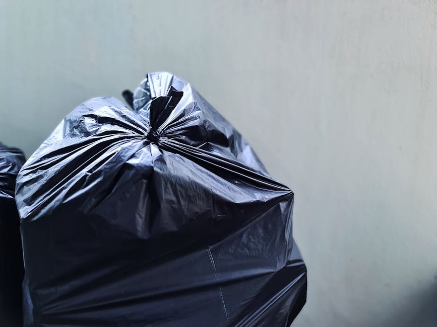 Close-up view of two large black rubbish bags made of glossy plastic, tightly sealed with twisted tops, positioned in the foreground against a plain, light-colored wall. The surface of the bags displa